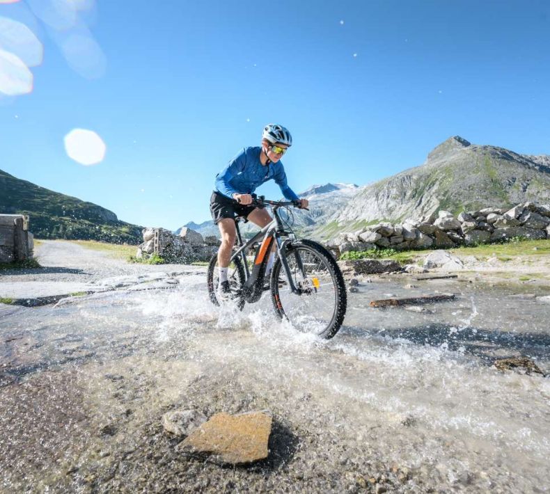 Ein Mountainbiker überquert einen Wasserlauf auf einem Pfad, umgeben von Berglandschaften