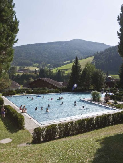 Ein Freibad mit Menschen, die im Wasser spielen, umgeben von grüner Landschaft und Bergen