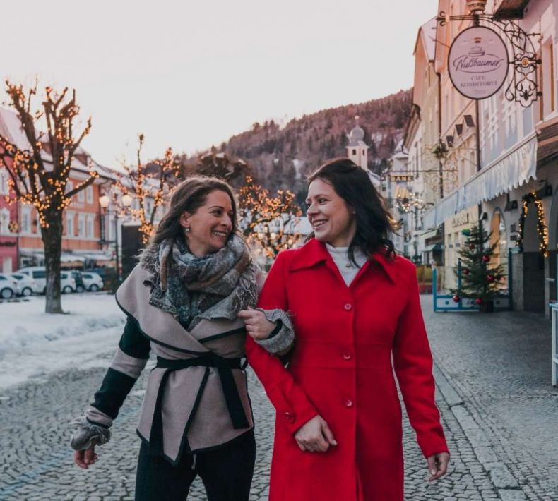 Zwei Frauen gehen lachend durch eine winterliche Stadt, geschmückt mit Weihnachtsbeleuchtung